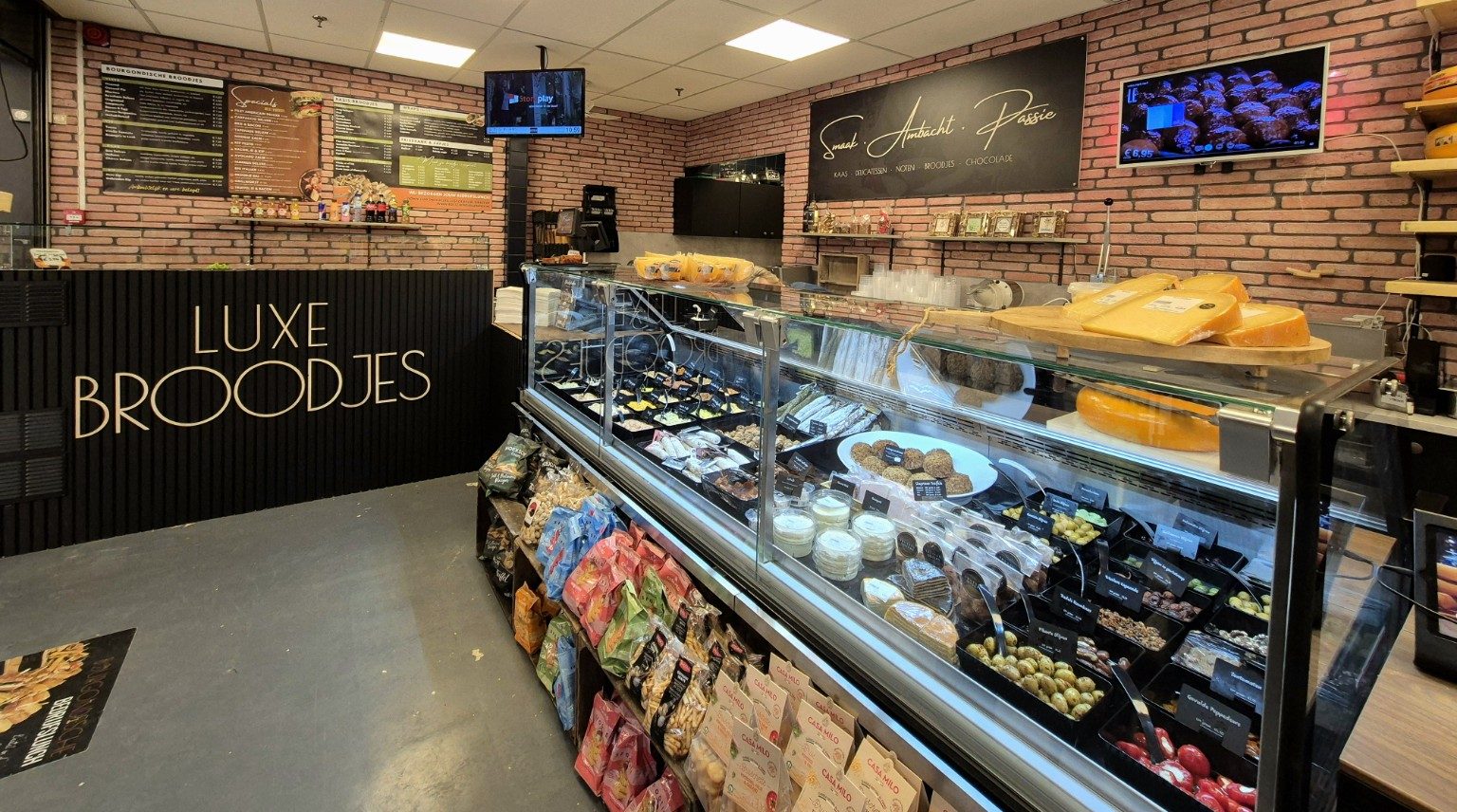 Onze winkel in Den Bosch: de toonbank met luxe broodjes, de vitrine vol kazen en delicatessen en de bakstenen wand met Smaak · Ambacht · Passie